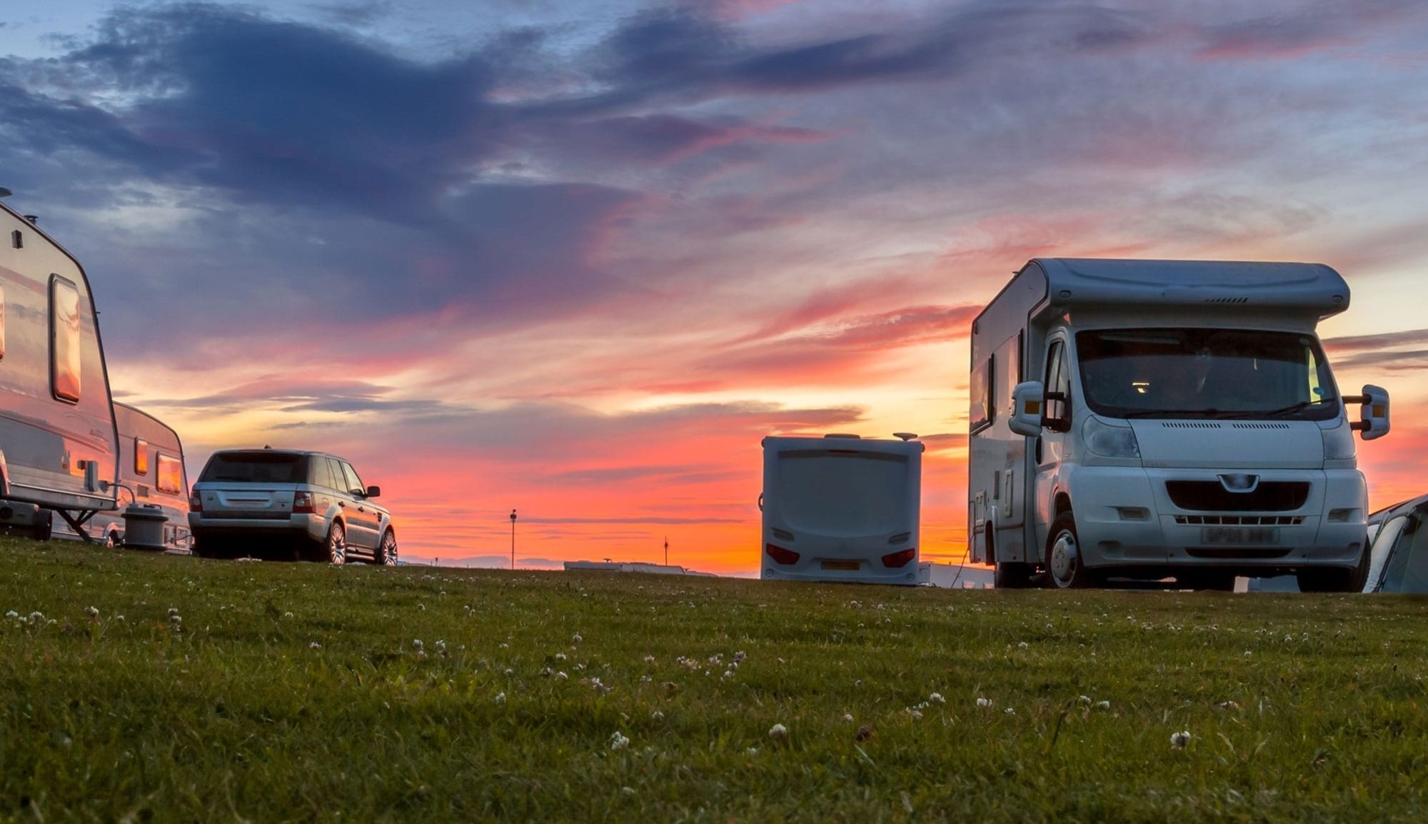 Campingvogn og autocamper parkeret på græs ved solnedgang med farverig himmel.